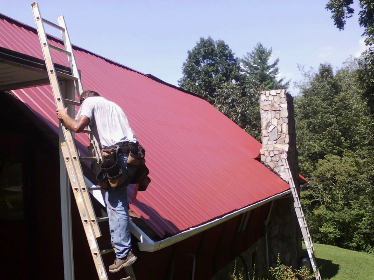 Roofer on a ladder beside a red metal roof and stone chimney — Soffit & Fascia Repair by Roofing Brothers in Glen Lyon